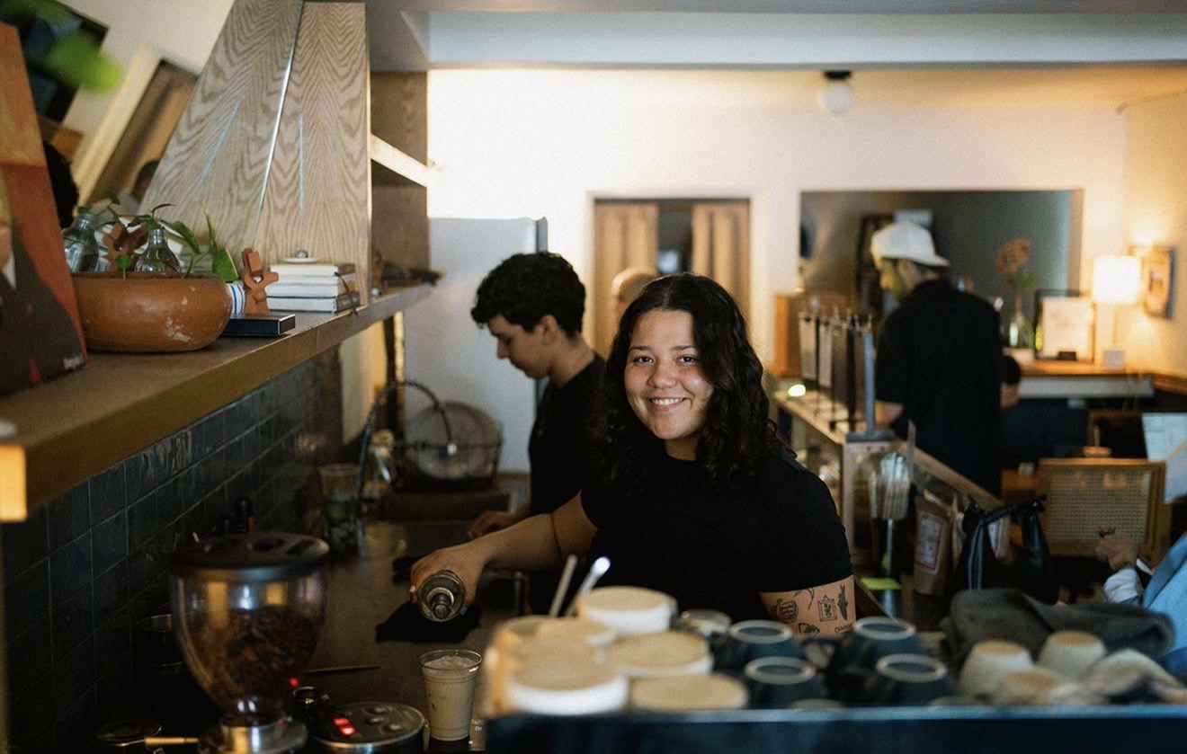 Barista sonriendo detrás de la barra de Josefina mientras prepara bebidas, con molino de café y tazas al frente