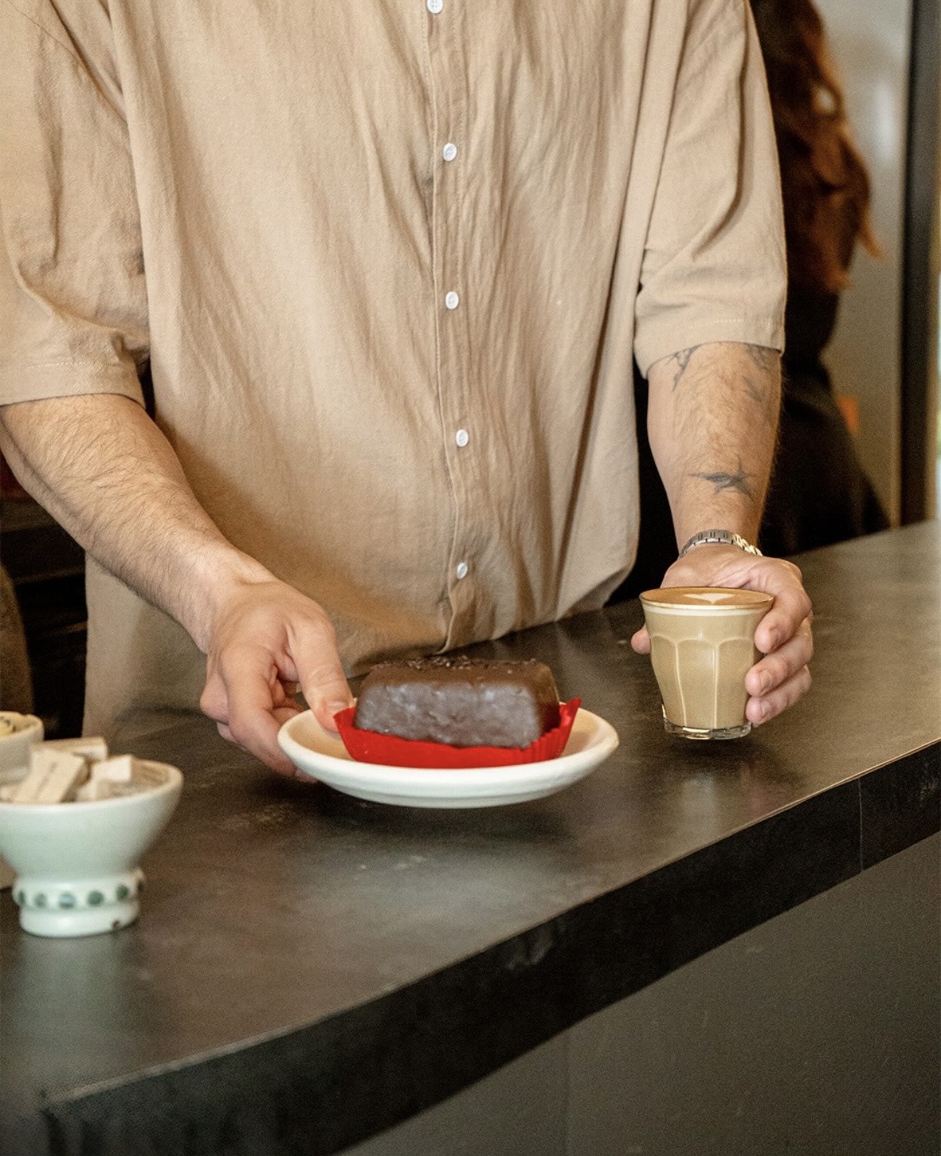 Persona colocando un pan de chocolate en un plato sobre la barra, con un vaso de café con leche en la mano