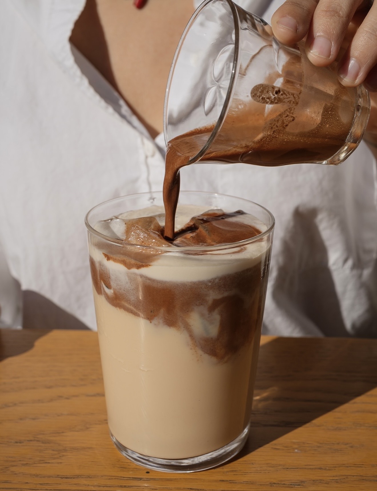 Cacao siendo vertido desde un vaso de vidrio sobre un vaso con leche y hielo, sobre una mesa de madera