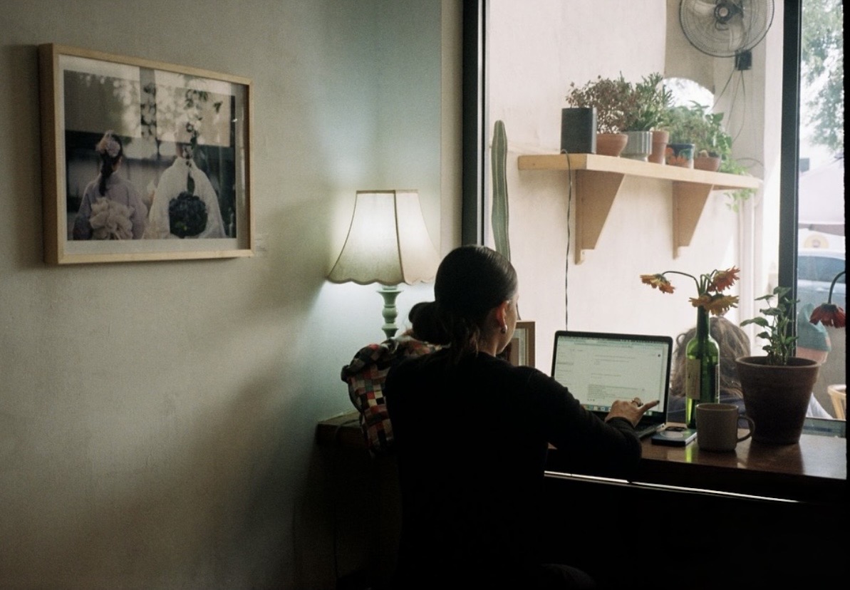 Interior de Josefina con una persona trabajando junto a la ventana, una lámpara encendida y plantas en la repisa