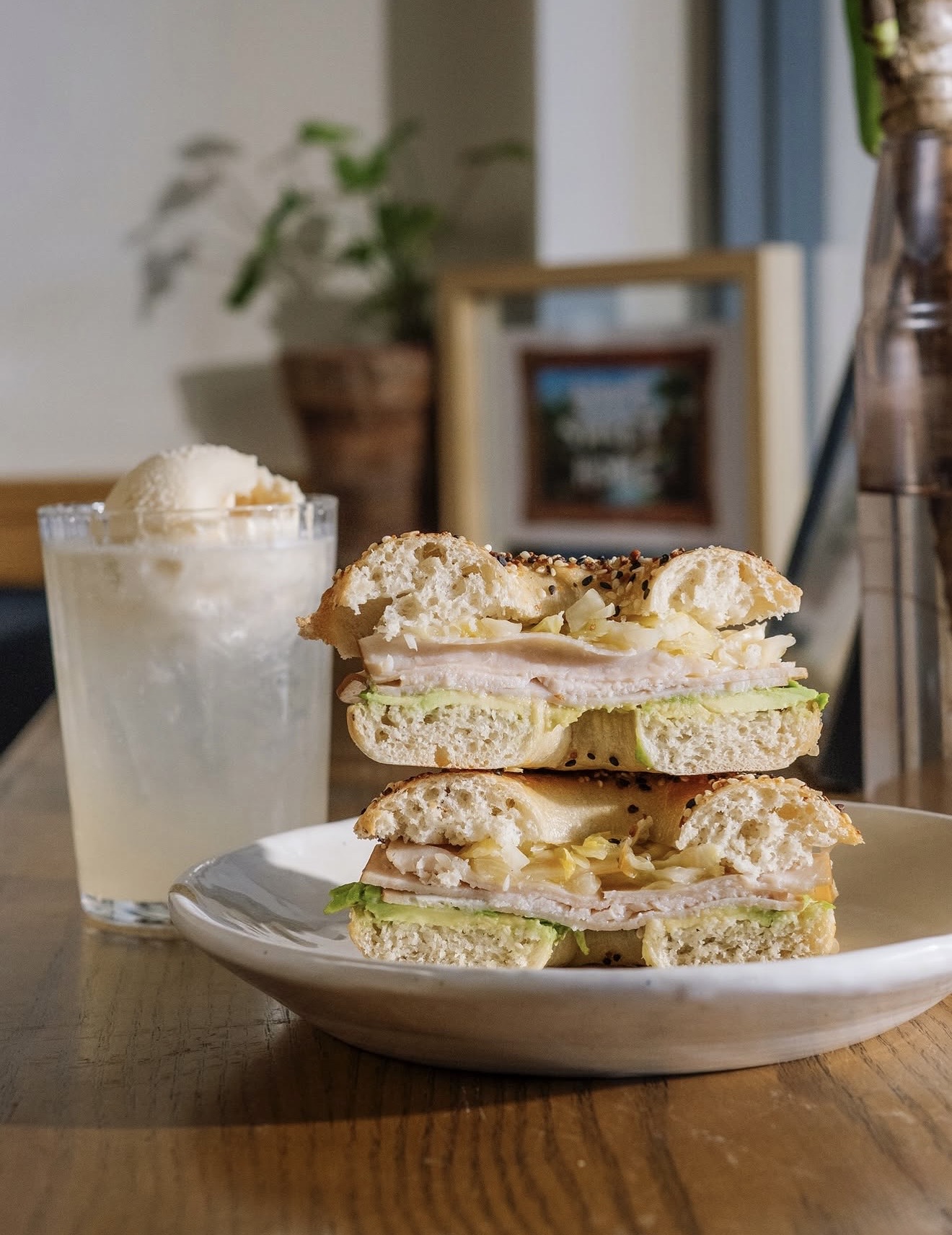 Bagel partido a la mitad con pavo, aguacate y chucrut en un plato blanco, junto a un vaso con hielo y una bola de helado