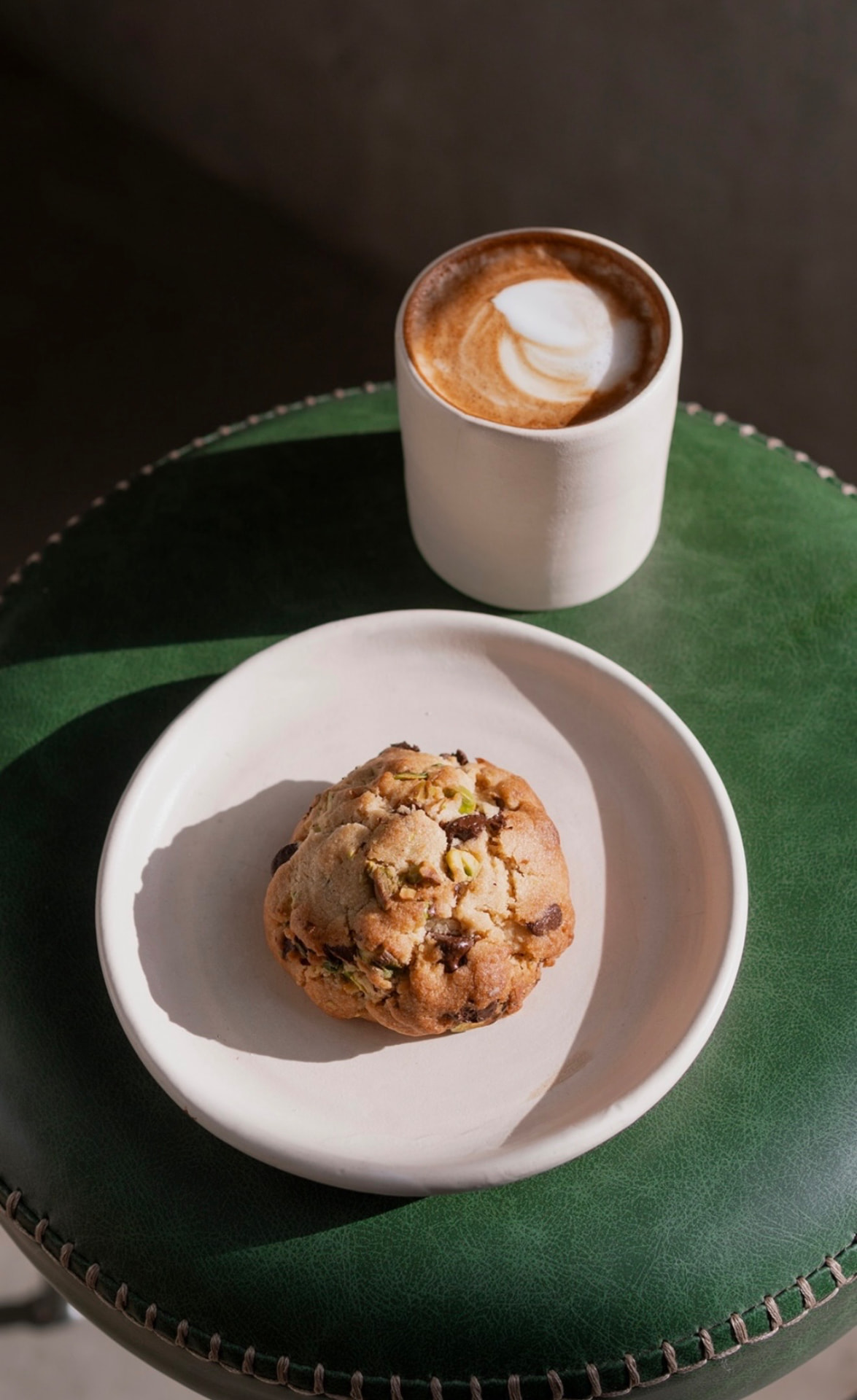 Galleta con pistache y chocolate en un plato blanco junto a un capuchino sobre un banco de piel verde